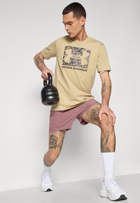Camiseta beige de Under Armour con un logotipo de camuflaje, combinada con pantalones cortos deportivos rosas, sosteniendo una kettlebell negra, zapatos deportivos blancos y calcetines.