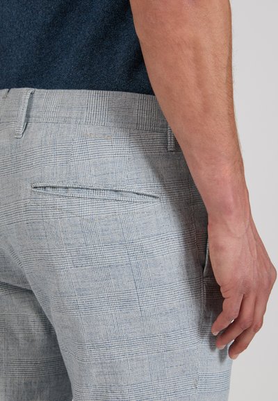 Man’s arm resting beside light gray plaid trousers with a back slit pocket and a dark blue shirt tucked in.
