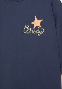Tissu bleu foncé avec un insigne de shérif et le mot "Woody" écrit en dessous dans une écriture ressemblant à une corde en haut à gauche.