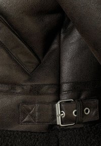 Veste en cuir marron avec une finition texturée, dotée d'une sangle et d'une boucle en métal. En dessous, il y a un matériau sombre et duveteux.