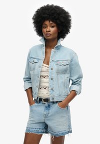 Chaqueta de mezclilla azul claro con cuello plegable y bolsillos en el pecho, combinada con una blusa de encaje blanca y pantalones cortos de mezclilla azul claro con bordes deshilachados.
