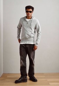 Gray zip-up hoodie with drawstrings, layered over a white shirt, paired with dark jeans and black shoes. Model wears black sunglasses.