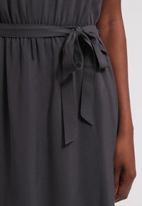 Robe gris foncé avec taille froncée cintrée par une ceinture en tissu nouée, portée par une personne à la carnation intermédiaire foncée.