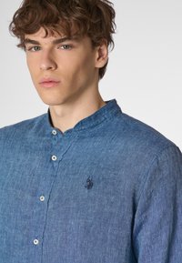 Camicia blu in chambray con colletto alla coreana, caratterizzata da un piccolo logo ricamato e bottoni bianchi, che mette in evidenza un tessuto testurizzato.