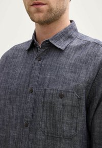 Cămașă cu nasturi din denim gri închis, având un guler rezistent, buzunar pe piept și nasturi maro. Textură netedă cu un model subtil.
