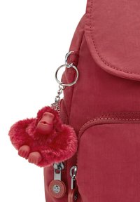 Mochila de tela roja que cuenta con un bolsillo frontal con cremallera, una solapa redondeada y un llavero de un mono rojo esponjoso sujeto por un anillo de plata.