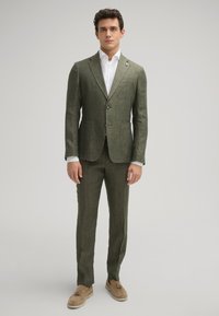 Olijfgroen linnen pak met een getailleerde pasvorm, met notch revers, twee knopen en klepzakken. Gepaard met een wit overhemd en beige instappers.