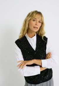 Veste noire avec des sequins, décolleté en V, boutons sur le devant et bords côtelés. Portée par-dessus une chemise blanche à manches longues et à coupe décontractée.