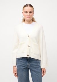 Kremno obarvan cardigan z V-izrezom, petimi gumbi in dvema sprednjima žepoma. Mehka tekstura in rebrasti manšeti ter spodnji del. Nosi se preko bele majice.