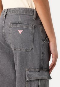 Cargo pants in dark gray denim, featuring a back pocket with a red and white triangular logo, and a side pocket with a flap.