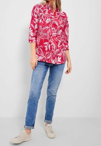 Femme portant une chemise à boutons rouge à fleurs, un jean bleu clair retroussé et des baskets blanches, se tenant devant un fond clair uni.