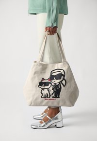 Kanvasas iepirkumu soma bēšā krāsā ar melnu karikatūru grafiku, kurā attēlots vīrietis un kaķis, abi valkā saulesbrilles. Iemaukti ir izturīgi, un audums ir mīksts.