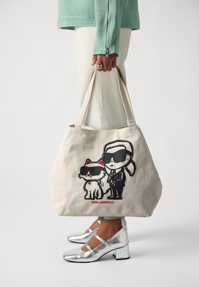 Kanvasas iepirkumu soma bēšā krāsā ar melnu karikatūru grafiku, kurā attēlots vīrietis un kaķis, abi valkā saulesbrilles. Iemaukti ir izturīgi, un audums ir mīksts.