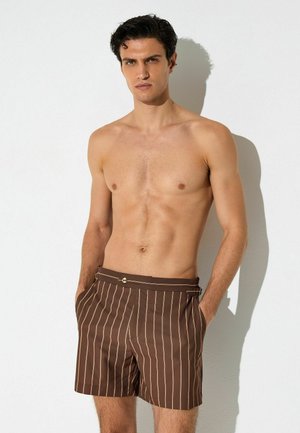 T-särgita mees, tume juustega, pruunide pükstega, millel on õhukesed vertikaalsed beežid triibud, seisab valge seina ees, käed taskutes.