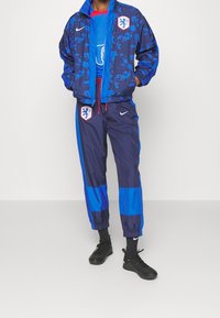 Blå Nike-träningsdress med mönstrad jacka, enfärgade blå detaljer, elastiska muddar och ett emblem på jackan och byxorna. Svarta sportskor.