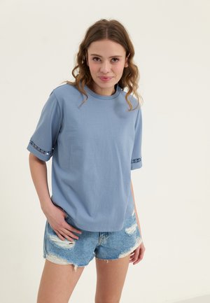 Lyseblå kortermet skjorte med rund hals og blonde detaljer på ermene, sammen med slitte denimshorts.