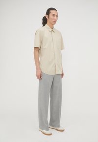 Beige kortleurige overhemd met knopen en een borstzak, gecombineerd met lichtgrijze wijde broekspijpen en witte sneakers met een bruine zool.