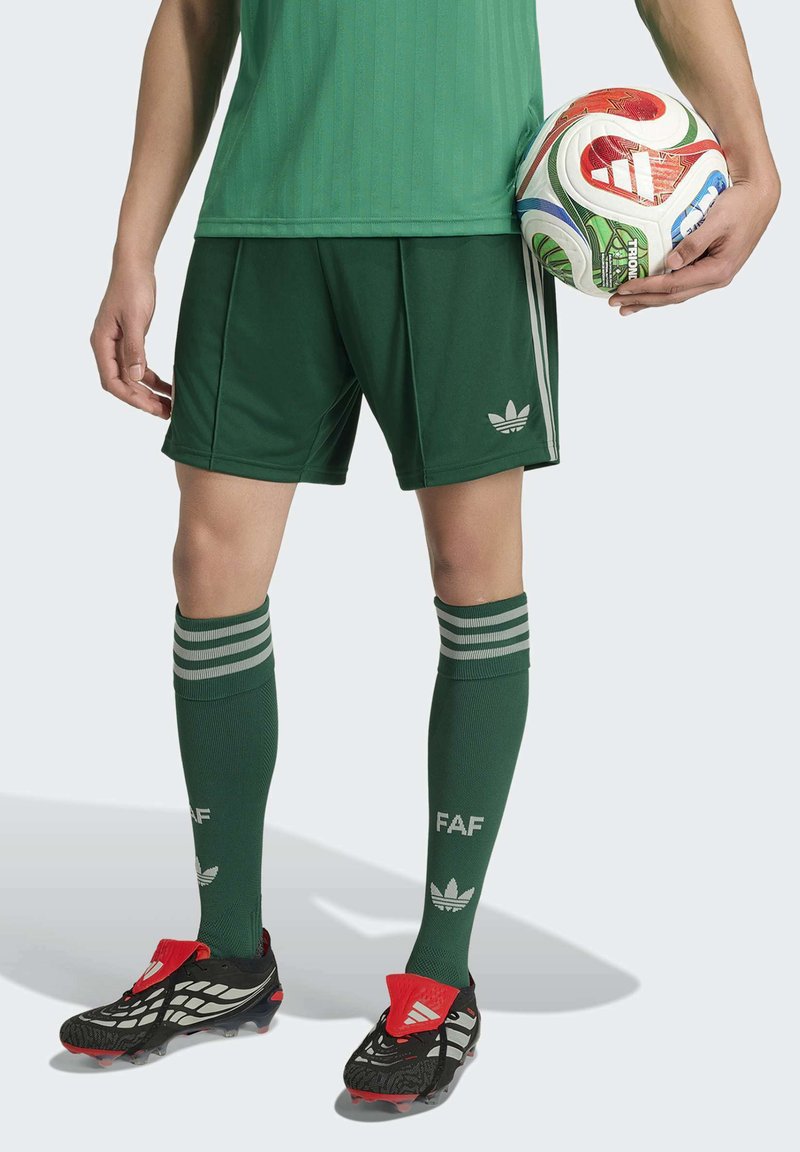 Personne portant un équipement de football vert et des crampons, tenant un ballon de football Adidas blanc, rouge et vert à côté d'elle.