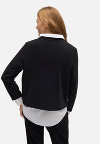 Chaqueta exterior negra y estructurada con mangas largas; presenta un dobladillo de camisa blanca superpuesto por debajo, proporcionando un contraste de color y textura.