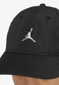 Černá baseballová čepice s texturovanou látkou, na přední straně bílý vyšívaný logo Jumpman, zahnutý kšilt a otvory pro ventilaci.