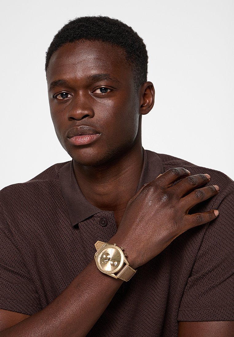 Montre bracelet en or avec un bracelet en maille texturée, dotée d'un cadran rond, de trois sous-cadrans et d'une finition polie. Portée sur une chemise polo marron.