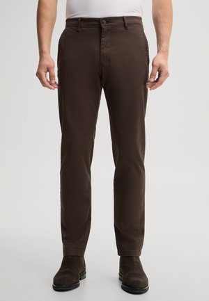 Mann trägt dunkelbraune gerade geschnittene Hose und dunkelbraune Wildlederstiefel, steht vor einfarbig hellem Hintergrund.