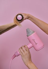 Vaaleanpunainen hiustenkuivaaja, jossa on harmaa suutin, pidetään yhdessä kädessä ja muotoillaan vaaleita hiuksia pyöreällä harjalla toisessa kädessä, vaaleanpunaisella taustalla.