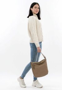 Pull beige en maille torsadée, jean skinny bleu clair et baskets blanches, portant un sac à bandoulière en cuir marron avec un détail en fermeture éclair.