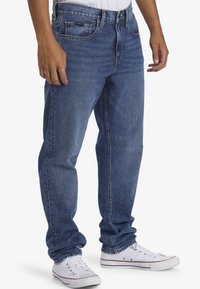 Modré denimové džínsy s voľným strihom, obsahujúce päť vreciek, zapínanie na gombík a zips a jemné detaily prešitia. Na nohe má biele tenisky.