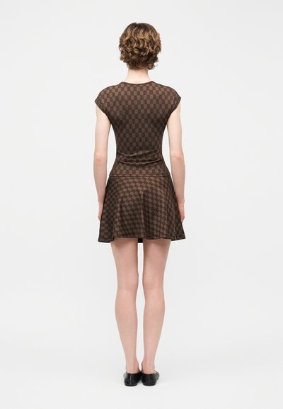 Γυναίκα που στέκεται με την πλάτη προς την κάμερα, φοράει καφέ κοντομάνικο πουκάμισο με σχέδια και τζιν φούστα με καμπάνα, μαζί με μαύρα παπούτσια σε απλό φόντο.