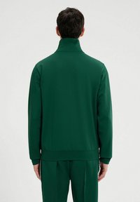 Chaqueta deportiva verde con cuello alto, material elástico, textura suave y dobladillo acanalado; combinada con pantalones a juego.