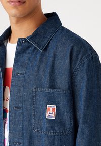 Tummansininen farkkupaita, jossa on kauluspääntie, rintatasku ja Wrangler-logo-ompelu. Kankaan pinta on teksturoitu.