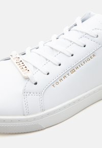 Baskets en cuir blanc avec lacets blancs, logo Tommy Hilfiger doré sur le côté, et une étiquette métallique dorée sur les lacets.