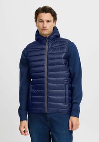 Jūras zilas pufēts vestes ar augstu apkakli, priekšējo rāvējslēdzēju un horizontālu quilted rakstu. Valkāta virs garām piedurknēm tumši zilas krāsas krekla.
