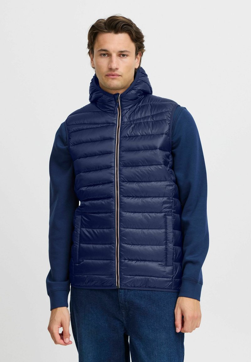Jūras zilas pufēts vestes ar augstu apkakli, priekšējo rāvējslēdzēju un horizontālu quilted rakstu. Valkāta virs garām piedurknēm tumši zilas krāsas krekla.