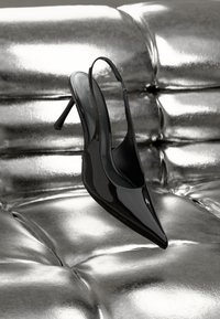 Čierne lakované kožené slingback podpätky s špicatou špičkou a tenkým stiletto podpätkom, vystavené na lesklom striebornom polstrovanom povrchu.