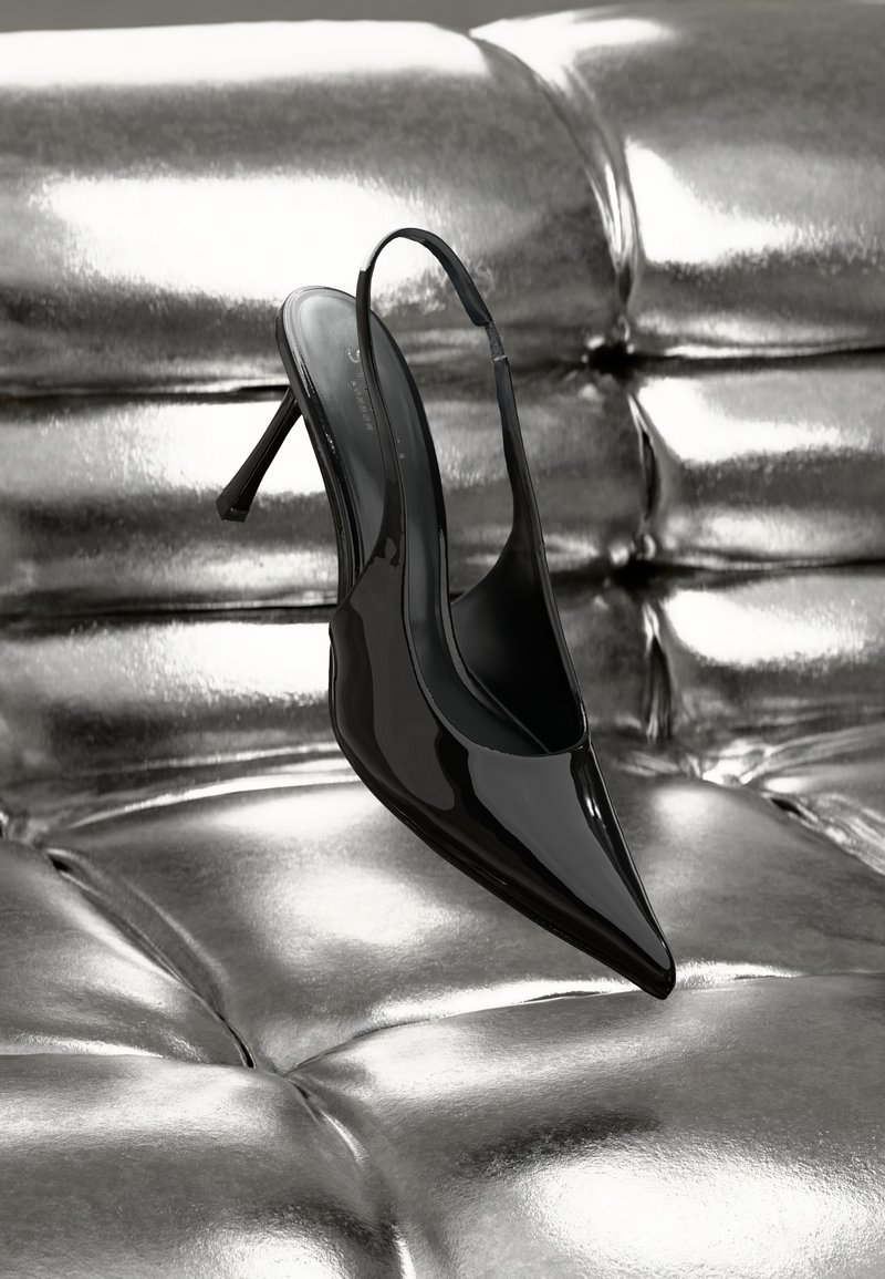 Čierne lakované kožené slingback podpätky s špicatou špičkou a tenkým stiletto podpätkom, vystavené na lesklom striebornom polstrovanom povrchu.