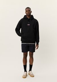 Μαύρο φούτερ με κουκούλα και τσέπη μπροστά, κορδόνι στην κουκούλα και μικρό έμβλημα λογότυπου. Συνδυάζεται με σκούρα σορτς και ανοιχτόχρωμα αθλητικά παπούτσια. Ομαλή υφή.