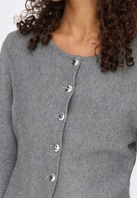 Femme portant un cardigan gris en maille avec des boutons ronds marbrés noirs et blancs, cheveux bouclés visibles autour du cou et des épaules.
