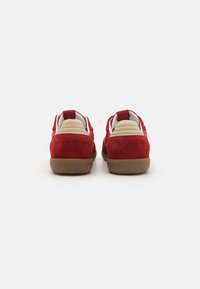 Baskets en suede rouge avec des accents beiges et des cols rembourrés, dotées d'une semelle en caoutchouc texturé et de languettes à l'arrière pour un enfilage facile.