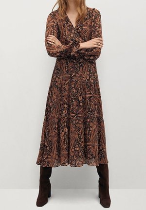 Žena v dlouhých šatech s hnědým paisley vzorem a černými a oranžovými květinovými detaily, stojící se založenýma rukama, obutá do tmavě hnědých bot.