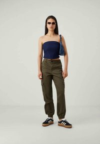 Jūrų karinio laivyno be petnešų viršus, alyviniai krovinių džinsai su elastiniais rankovėmis, juodi sportbačiai su baltais dryžiais ir saulės akiniai. Priedai – mėlynas krepšys.