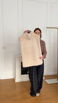 Jersey oversize de color beige claro con un cuello alto y doblado. Tejido suave y texturizado. La modelo lo sostiene de una percha contra un fondo liso.