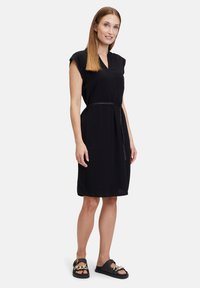 Schwarzes knielanges Kleid mit kurzen Ärmeln, Rundhalsausschnitt und einem Stoffgürtel in der Taille; glatte Textur. Kombiniert mit schwarzen Sandalen mit Kettenakzent.