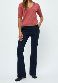 Czerwony sweter z krótkim rękawem w serek, zestawiony z granatowymi spodniami w dzwony. Ubranie charakteryzuje się dopasowanym krojem oraz eleganckimi czarnymi butami.