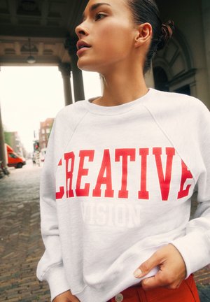 Femme aux cheveux en chignon porte un sweat-shirt blanc avec un grand texte rouge "CREATIVE", debout dans une rue pavée près de colonnes architecturales.