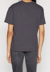 Camiseta de algodón gris con mangas cortas, cuello redondo y un corte relajado. Detalles de costura a lo largo del dobladillo. Llevada con jeans gris oscuro.