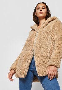 Vaahtokarkki beige fleece-takki hupulla, jossa on vetoketjukiinnitys ja röpelöinen kangas. Lievästi löysä ja pehmeä, leveillä hihoilla ja hieman pidemmällä helmalla.