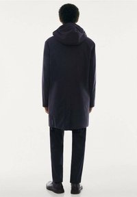 Abrigo largo de color azul marino oscuro con capucha, corte recto y una textura sutil. Llevado sobre pantalones oscuros y combinado con zapatos negros informales.