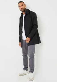 Abrigo impermeable negro con cuello, cinco botones y un diseño aerodinámico, combinado con pantalones grises y zapatillas blancas.